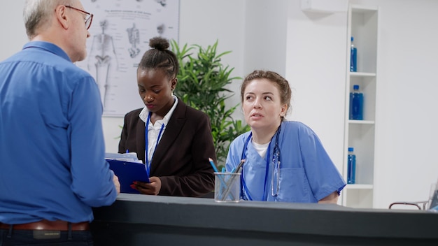 Equipo médico diverso que trabaja en la recepción del hospital, ayudando a los pacientes con citas de control y documentos de informes. Enfermera y recepcionista sentadas en el mostrador de registro en el vestíbulo de las instalaciones.