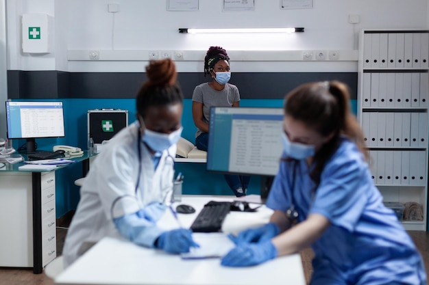 Equipo médico con mascarilla protectora para prevenir infecciones con covid19 analizando documentos de medicación trabajando en la oficina del hospital. Médico afroamericano que examina al paciente enfermo durante la consulta