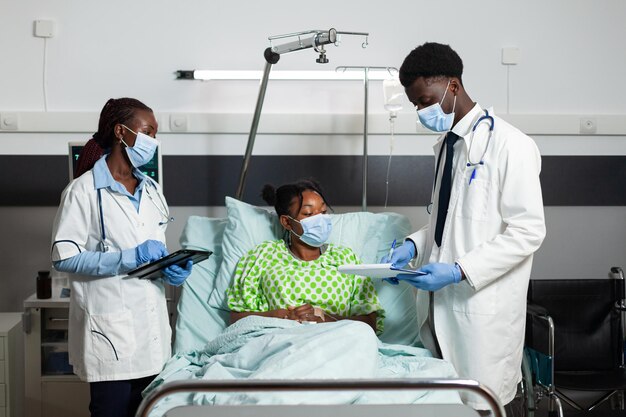 Equipo médico con mascarillas protectoras para prevenir la infección con covid19 monitoreando al paciente enfermo durante la cita clínica en la sala del hospital. Doctores afroamericanos explicando el tratamiento sanitario