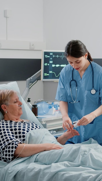 Equipo médico que realiza una visita de control para un paciente anciano con enfermedad en la cama de la sala del hospital. Médico y enfermera consultando a una anciana con oxímetro para saturación de oxígeno y bolsa de goteo IV.