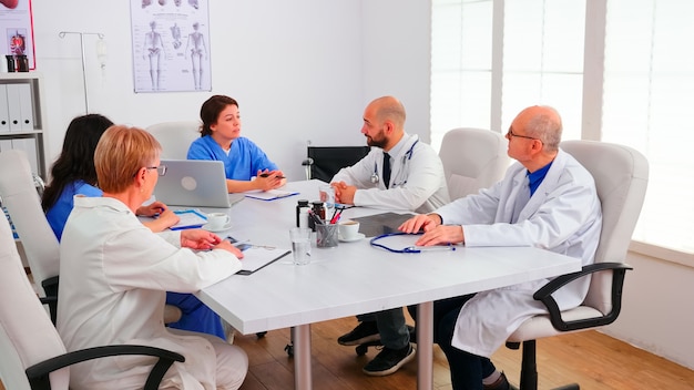 Equipo de médicos expertos sentados en el escritorio en la sala de conferencias del hospital con una sesión informativa. Terapeuta experto de la clínica hablando con colegas sobre la enfermedad para el desarrollo del tratamiento, profesional de la medicina