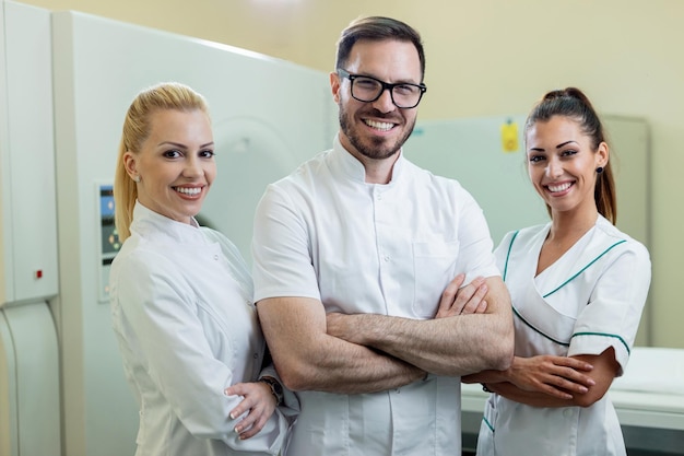 Equipo de médicos felices de pie con los brazos cruzados junto al escáner de resonancia magnética en el hospital y mirando a la cámara