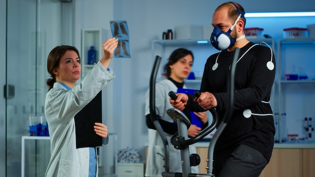 Equipo de médicos investigadores que supervisan la resistencia de los deportes de rendimiento del hombre con máscara que corre elíptica