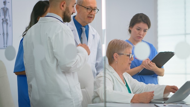 Equipo de médicos de pie en la sala de conferencias del hospital, médico senior discutiendo sobre el tratamiento del paciente en la computadora portátil. Compañeros de trabajo en batas blancas trabajando juntos analizando los síntomas de la enfermedad