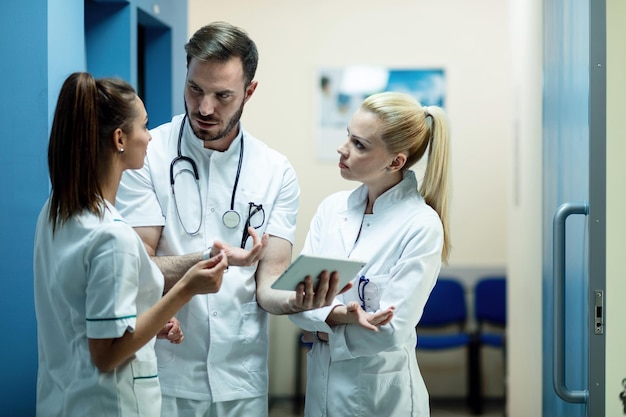 Equipo de médicos revisando informes médicos electrónicos en el panel táctil y hablando en un pasillo de la clínica