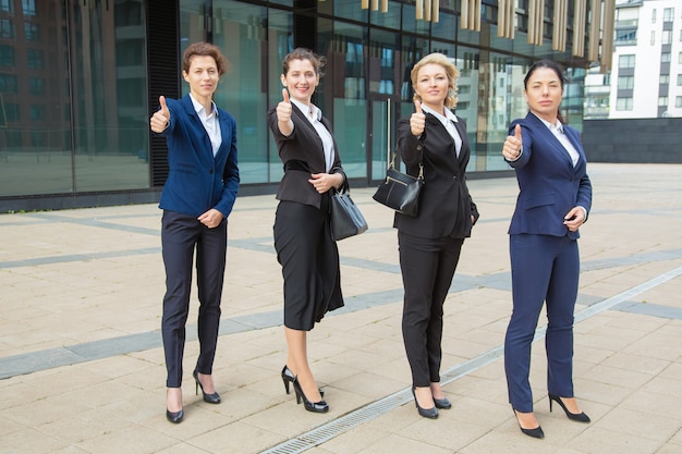 Equipo de mujeres empresarias de confianza positiva de pie junto al edificio de oficinas, mostrando como gesto, haciendo pulgar hacia arriba, mirando a la cámara. Longitud total. Concepto de éxito empresarial y trabajo en equipo