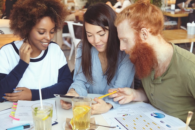 Equipo multiétnico de jóvenes emprendedores ambiciosos que trabajan juntos en la puesta en marcha del proyecto en la cafetería en la mesa con papeles de diagramas.