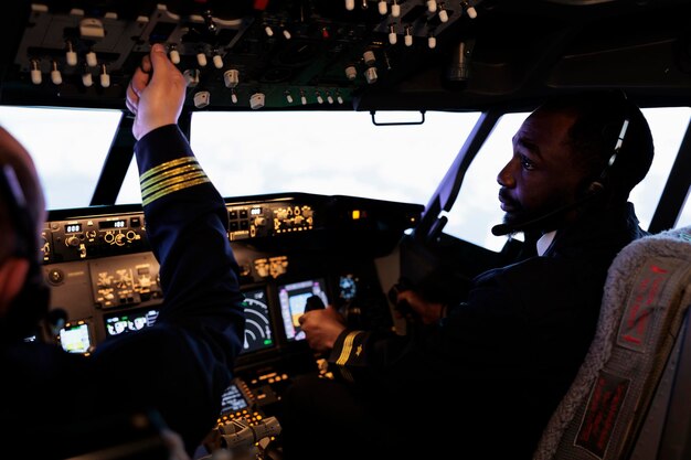 Equipo multiétnico de pilotos que usan el panel de control en la cabina para volar el avión, presionando el interruptor de encendido y los botones de comando del tablero para despegar. Servicio de vías aéreas internacionales con tripulación en cabina.