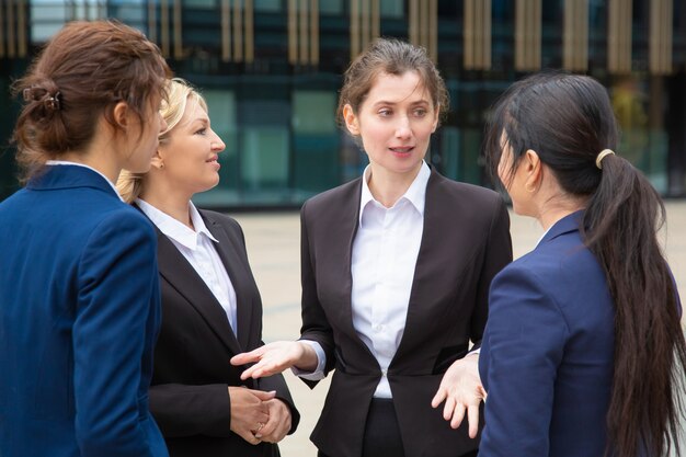 Equipo de negocios femenino creativo discutiendo el proyecto al aire libre. Empresarias con trajes de pie juntos en la ciudad y hablando.