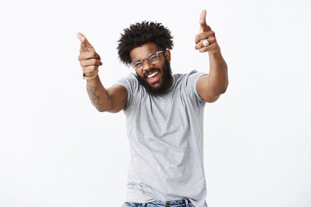 Eres un hombre de rock. Retrato de hombre alegre, guapo, emotivo, de piel oscura con peinado afro y barba apuntando con pistolas con el dedo a la cámara con las manos levantadas y una amplia sonrisa mientras le gustan los movimientos de baile de un amigo