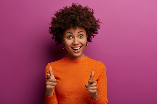 Eres lo que necesito. la mujer positiva y rizada te señala, te mira con alegría, quiere que te unas a su equipo, sonríe agradablemente, viste un poloneck naranja, posa sobre una pared morada, indica al frente