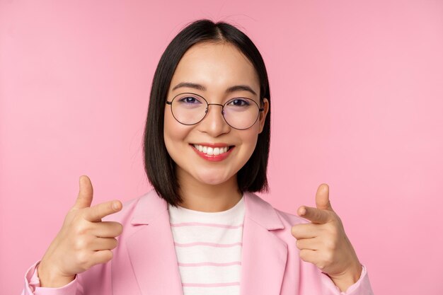 Eres tú, felicitaciones Sonriente directora ejecutiva asiática de mujer corporativa con traje y anteojos señalando con el dedo a la cámara reclutando elogios o cumplidos de pie sobre un fondo rosa