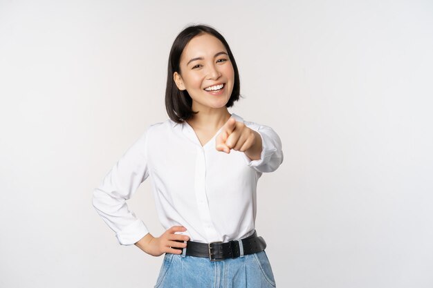 Eres tú Hermosa joven gerente de la empresa asiática señalando con el dedo a la cámara y sonriendo eligiendo invitar a la gente a reclutar de pie sobre fondo blanco