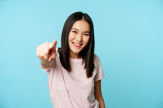Eres tu. Sonriente mujer asiática feliz apuntando con el dedo a la cámara, felicitando, invitando a la gente, de pie en camiseta sobre fondo azul.