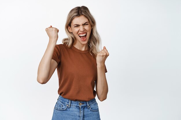 Ese es el espíritu. Entusiasta chica rubia ganando, celebrando la victoria, gritando de emoción y triunfo, de pie sobre blanco