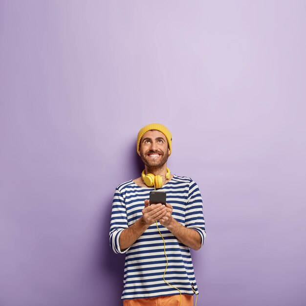 Es hora de escuchar música agradable. Alegre hombre caucásico tiene teléfono celular, enfocado hacia arriba en el espacio en blanco, disfruta de canciones geniales