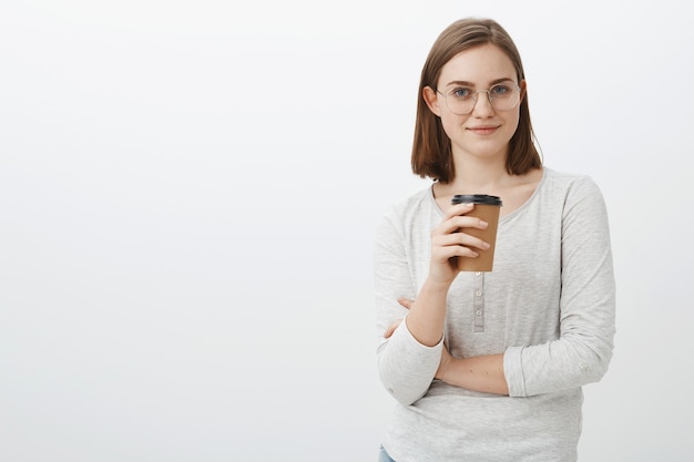 Es hora de tomar un café para trabajar productivamente. Satisfecha, relajada, alegre y atractiva mujer con gafas y blusa sosteniendo un vaso de papel de bebida y sonriendo burlonamente teniendo un descanso visitando un café hipster sobre una pared gris