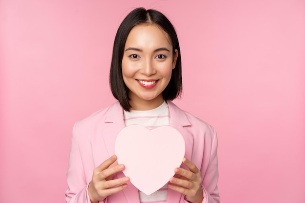 Esto es para ti Romántica linda mujer corporativa asiática con traje que muestra una caja en forma de corazón con un regalo de pie sobre un fondo rosa