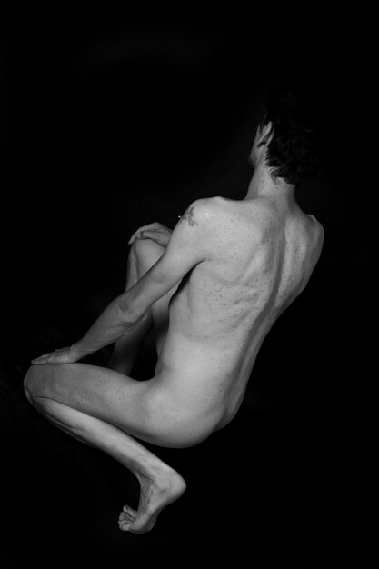 Escala de grises vertical de un hombre desnudo sentado en el suelo aislado en negro