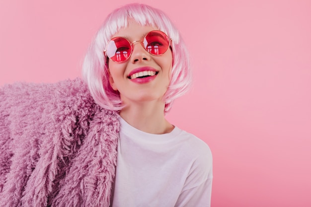 Escalofriante chica caucásica posando en gafas de sol y moda peruke. Debonair joven con cabello rosado riendo