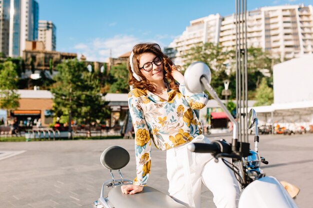 Escalofriante señorita en blusa vintage con estampado de flores sentado en ciclomotor y escucha música con árboles y rascacielos