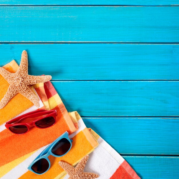 Escena de playa con toalla de rayas naranjas, estrellas de mar y gafas de sol en una vieja terraza de madera pintada de azul. Espacio para copia.