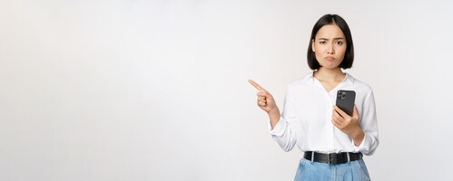 Foto gratuita la escéptica chica asiática moderna que sostiene el teléfono inteligente apuntando con el dedo a la izquierda en el espacio vacío y frunciendo el ceño tiene dudas sobre el fondo blanco