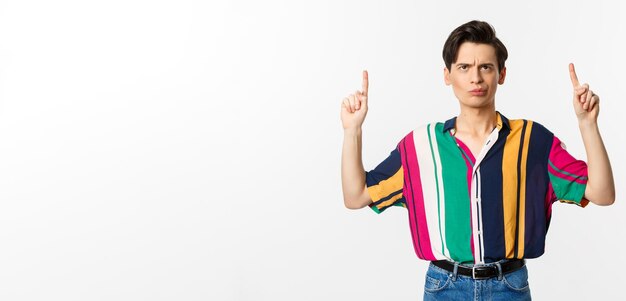 Escéptico y dudoso joven gay sonriendo señalando y mirando hacia la promoción vacilando de pie o