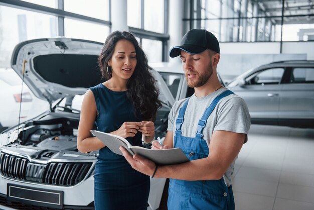 Escribir con la pluma. Mujer en el salón del automóvil con empleado en uniforme azul tomando su auto reparado