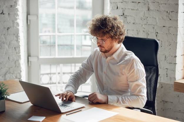 Escribir texto, analizar, navegar. Hombre joven caucásico en traje de negocios trabajando en oficina. Joven empresaria, gerente haciendo tareas con teléfono inteligente, computadora portátil, tableta tiene conferencia en línea, estudiando.