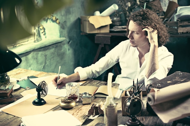 Foto gratuita escritor en el trabajo. apuesto joven escritor sentado en la mesa y escribiendo algo en su bloc de dibujo