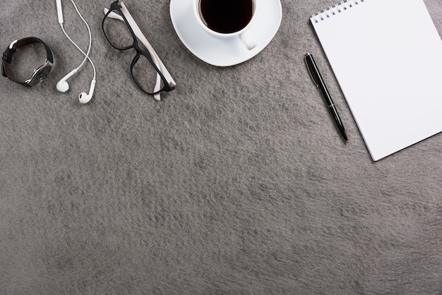 Escritorio de oficina gris con auricular; los anteojos; reloj de pulsera; taza de café; bolígrafo y cuaderno de espiral en blanco
