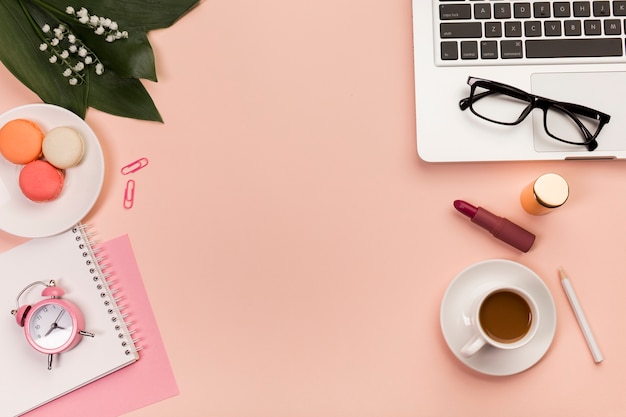 Foto gratuita escritorio de oficina con macarrones, taza de café, producto de maquillaje, lentes en la computadora portátil sobre el fondo de durazno