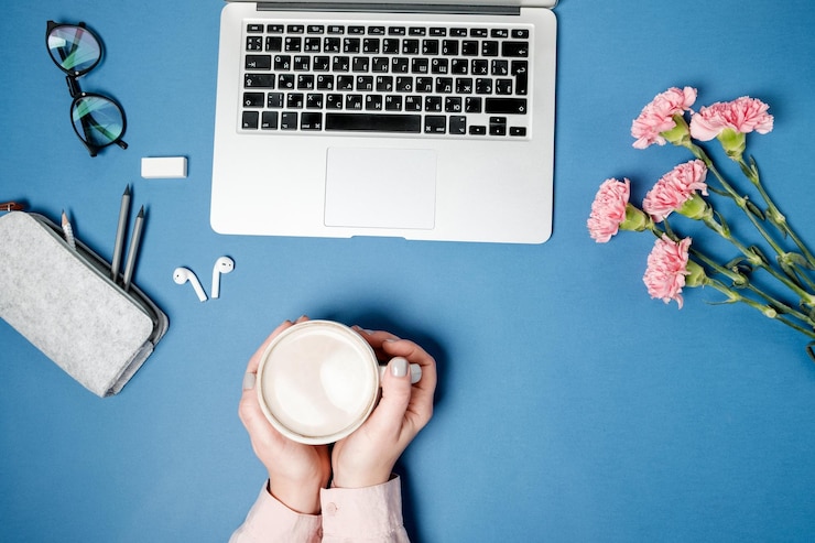 Foto gratuito escritorio de oficina plano para mujer manos femeninas con café y papelería de clavel rosa portátil en la mesa azul vista superior
