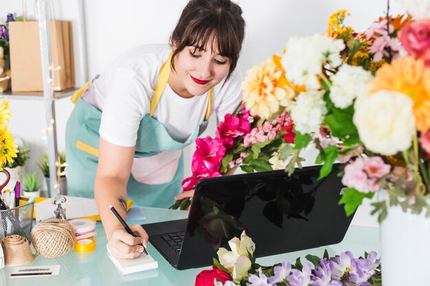 Escritura femenina del florista en la libreta con el ordenador portátil sobre el escritorio