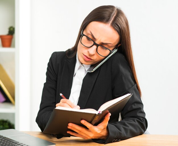 Escritura joven de la empresaria en el diario con el lápiz mientras que habla en smartphone