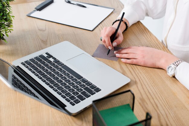 Escritura joven de la empresaria en nota con la pluma sobre el escritorio de madera con el ordenador portátil