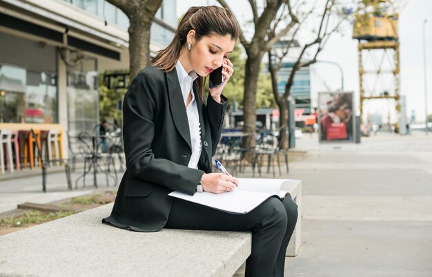 Escritura joven hermosa de la empresaria en carpeta con la pluma mientras que habla en el teléfono móvil