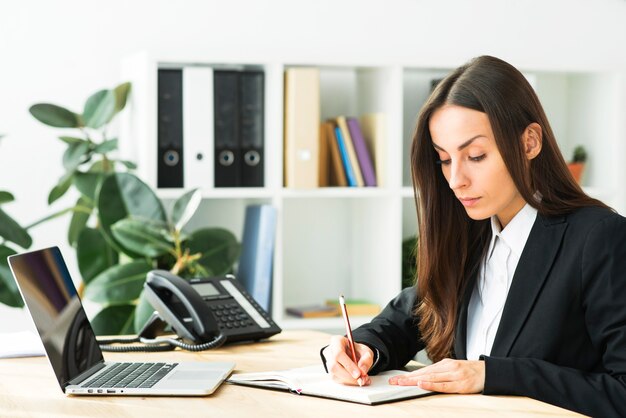 Escritura joven hermosa de la empresaria con el lápiz en el diario con el ordenador portátil sobre el escritorio