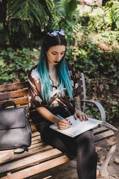 Escritura de la mujer joven en el cuaderno que se sienta en el banco en el parque