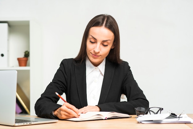 Escritura de la mujer joven en el diario con el lápiz en el lugar de trabajo en la oficina