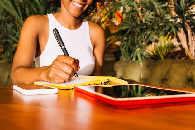 Escritura de la mujer joven en el diario con la pluma sobre la tabla de madera con el teléfono móvil y la tableta digital