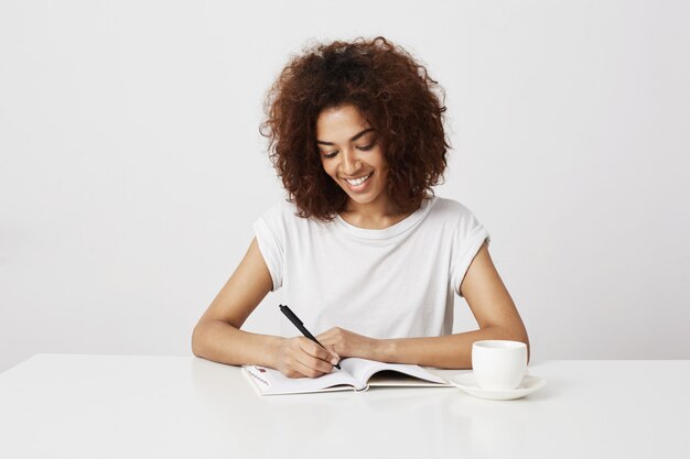 Escritura sonriente alegre de la muchacha africana en cuaderno en el lugar de trabajo sobre la pared blanca.