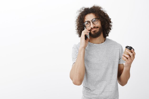 Escucha hombre, estoy ocupado. Retrato de chico hispano guapo fresco en gafas de moda y camiseta a rayas, sosteniendo la taza con café y hablando por teléfono inteligente