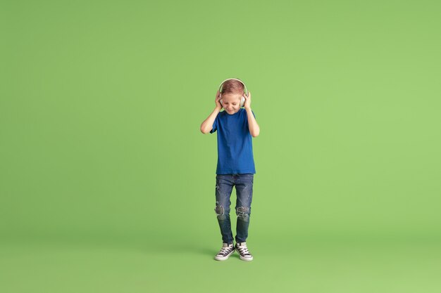 Escuche musi Happy boy jugando y divirtiéndose en la pared verde. Niño caucásico de aspecto brillante juguetón, riendo, sonriendo. Concepto de educación, infancia, emociones, expresión facial.