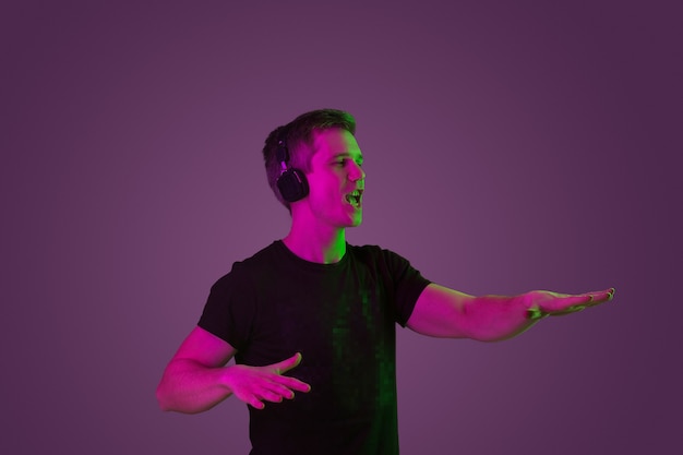 Escuche música, cante, disfrute. Retrato de hombre caucásico sobre fondo de estudio púrpura en luz de neón. Hermoso modelo masculino en camisa negra. Concepto de emociones humanas, expresión facial, ventas, publicidad.