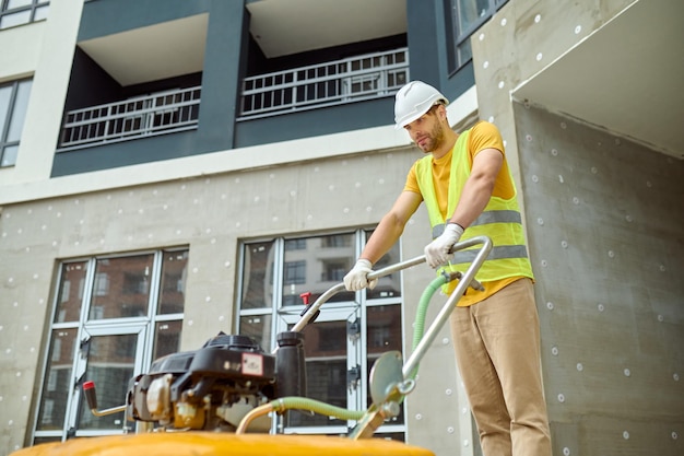 Esfuerzo, fatiga. Hombre cansado adulto joven en casco protector y chaleco brillante con equipo especial que trabaja en el sitio de construcción en el contexto del nuevo edificio