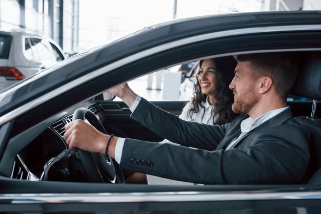 Eso luce bien. Encantadora pareja exitosa probando coche nuevo en el salón del automóvil