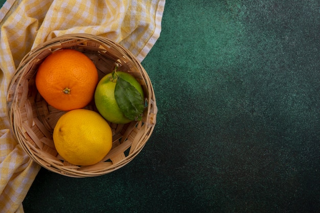 Espacio de copia de vista superior naranja con limón y lima en canasta con toalla a cuadros amarilla sobre fondo verde