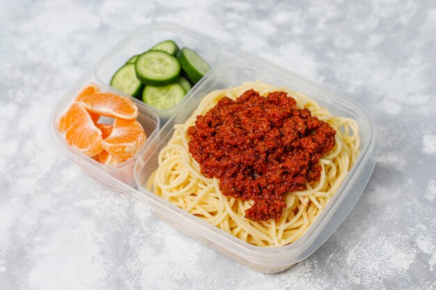 Espaguetis a la boloñesa para llevar en una lonchera de plástico con bebida de desintoxicación y una rodaja de fruta a la luz